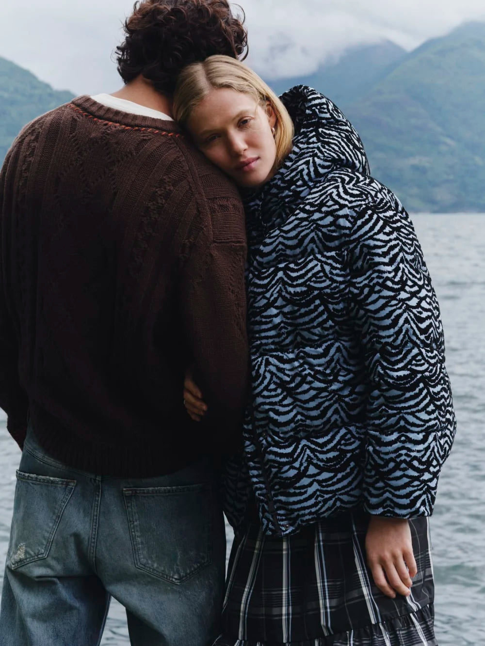 Two people standing close together by a body of water with mountains in the background.