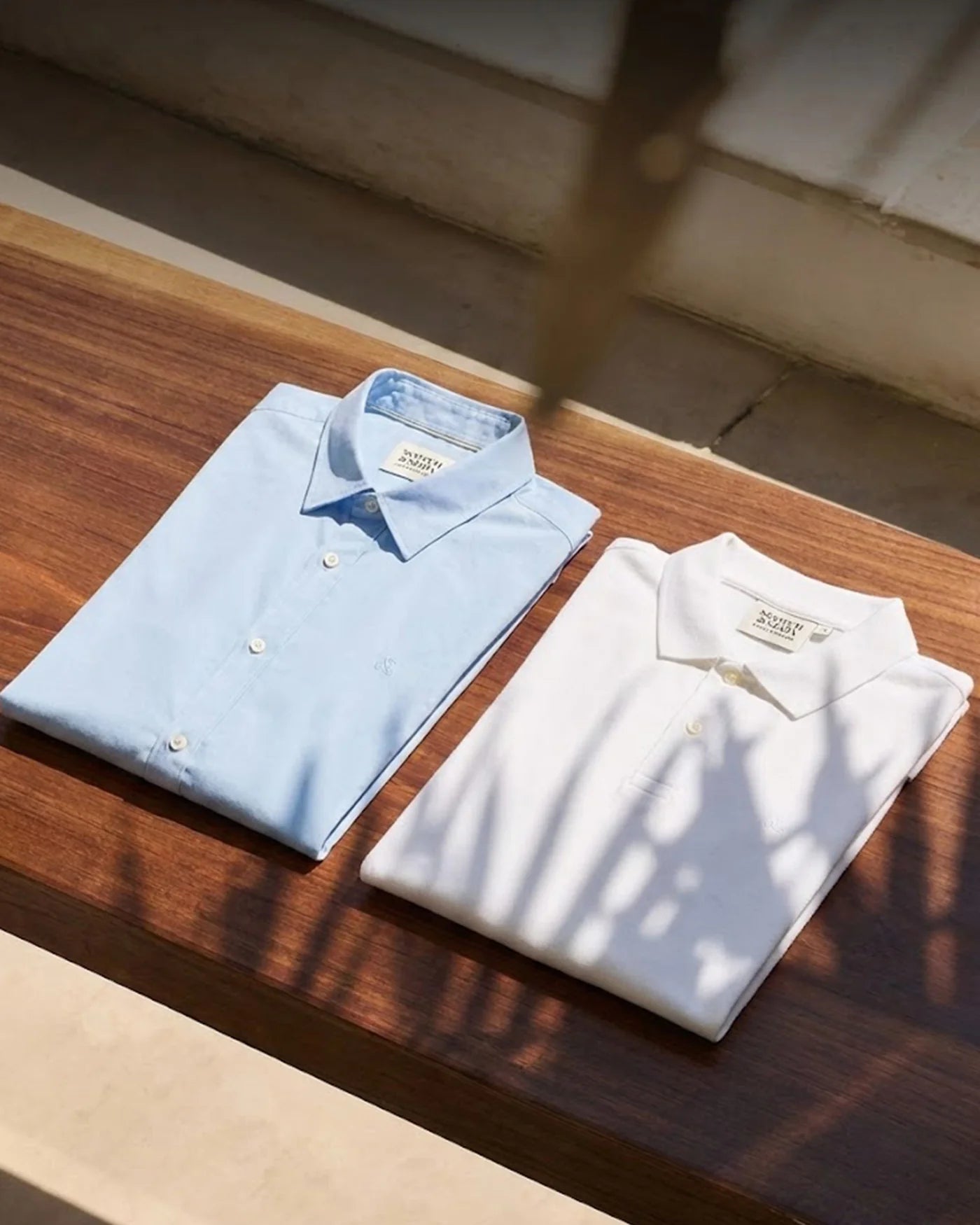 Two folded shirts, one blue and one white, on a wooden surface with sunlight casting shadows. Scotch Soda UK