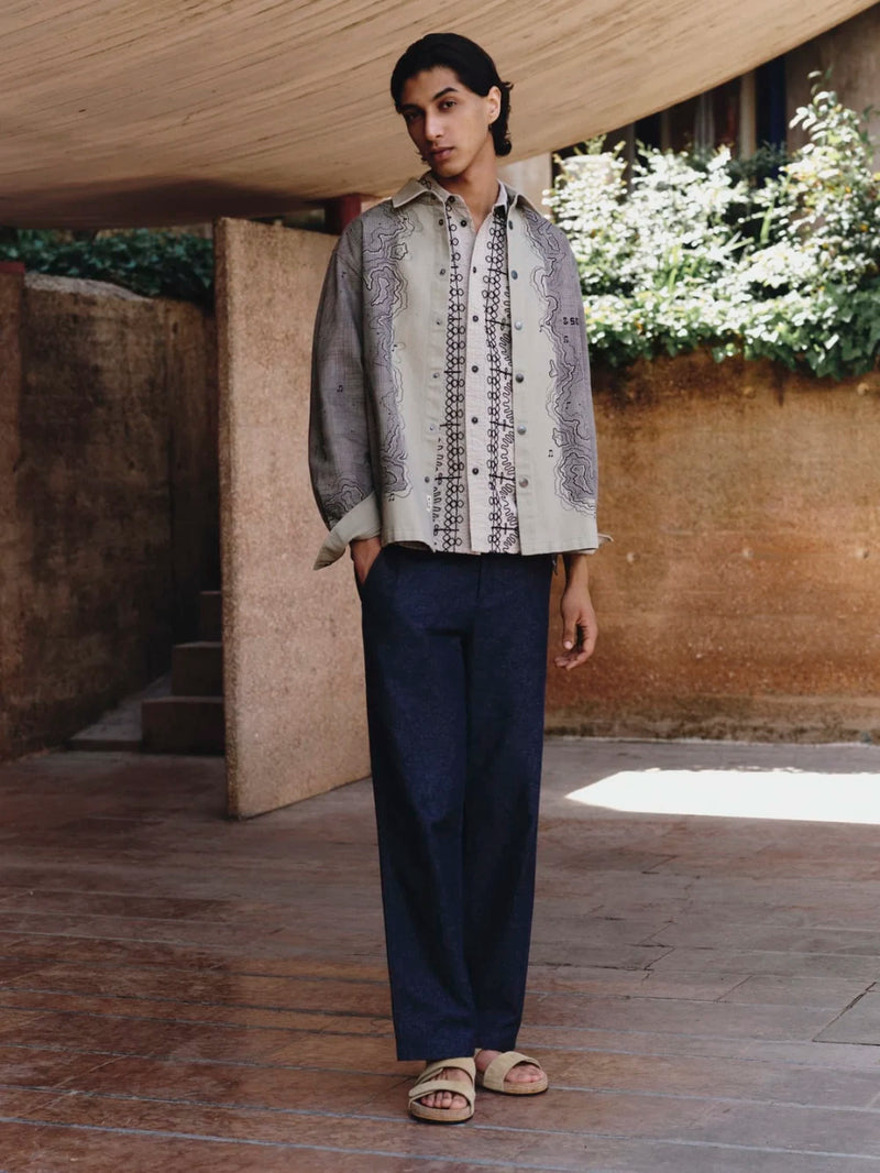Man wearing a patterned shirt and dark pants standing outdoors with greenery in the background