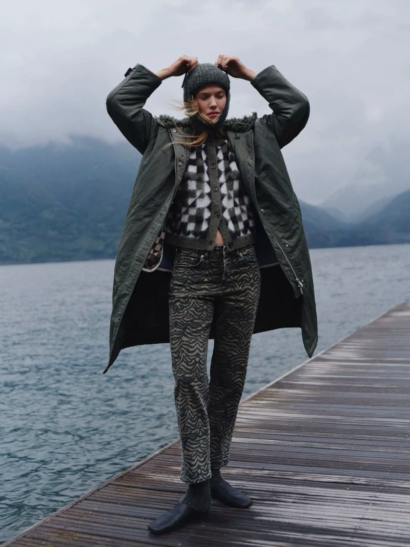 Person standing on a wooden dock by a lake, wearing a green coat and patterned pants.